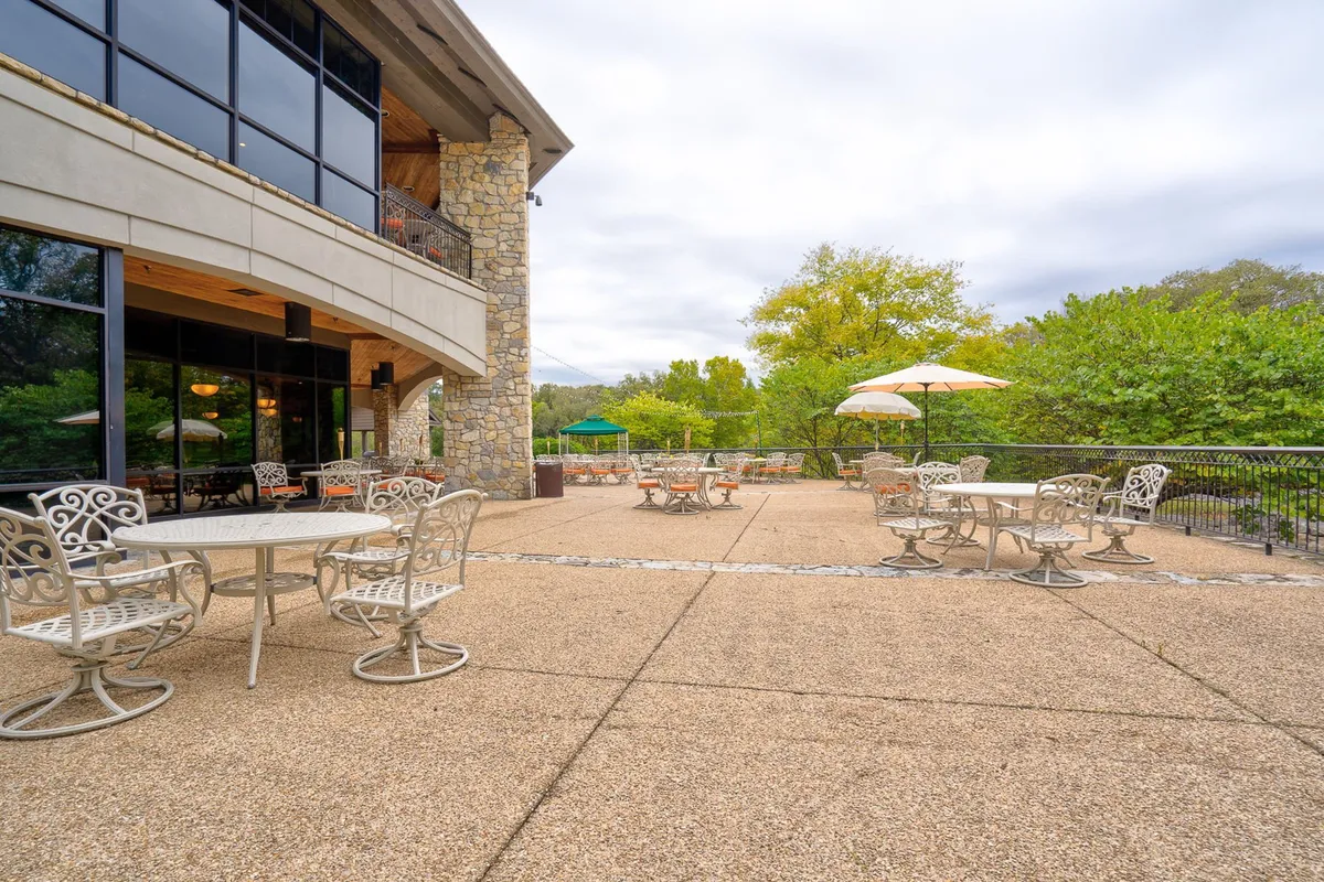 An exterior shot of a charming outdoor seating area with tables under umbrellas, large windows looking into the clubhouse or restaurant, and lush green trees beyond. The setup suggests al fresco dining, casual socializing, and a seamless connection to nature.