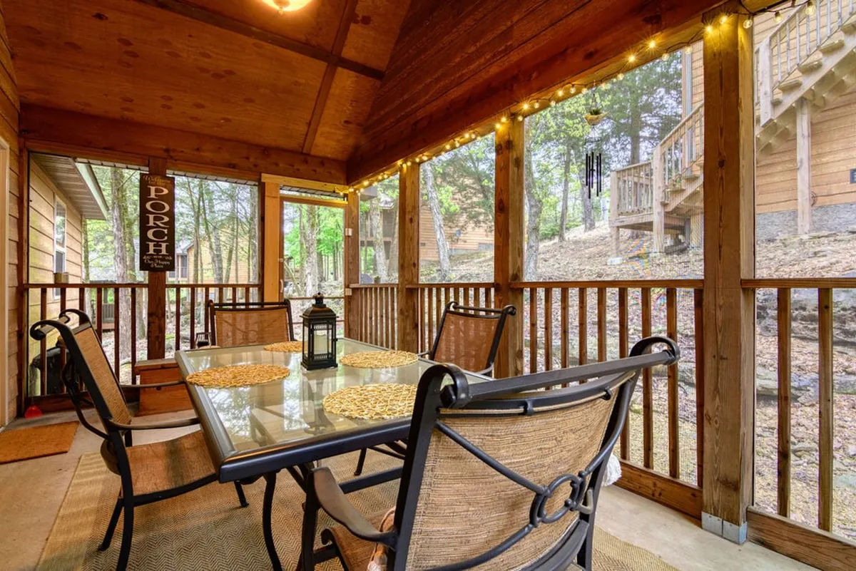 A warm, inviting dining space inside a cozy cabin, featuring a glass-top table set with woven placemats, a decorative lantern centerpiece, rustic wooden beams overhead, and twinkling string lights that create a relaxed, homelike ambiance.