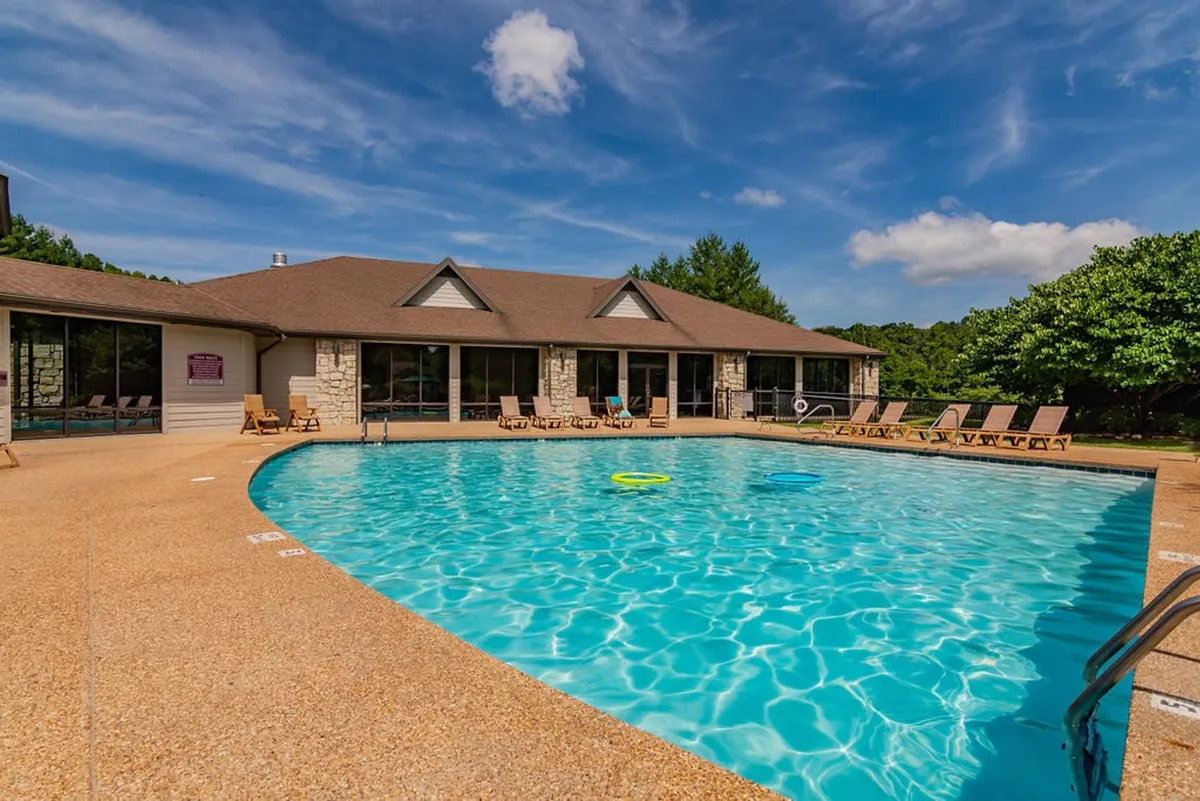 A tranquil pool area featuring inviting lounge chairs, a sparkling turquoise swimming pool, and a backdrop of resort architecture with large windows. The surrounding greenery and relaxed layout encourage socializing and unwinding after a day of activities.