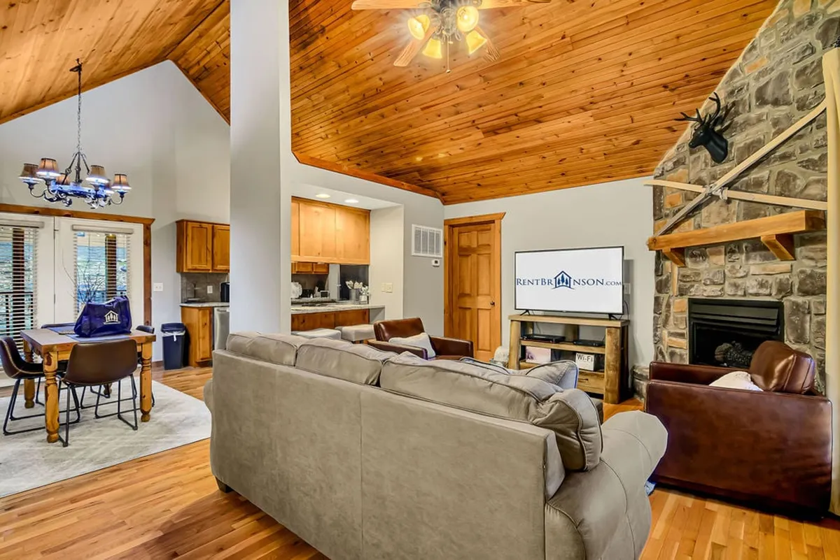 A wide-angle view of the open-concept living and dining area: the stone fireplace seamlessly connects to a wooden-ceilinged space with a comfortable seating area and a family-style dining table. The rustic décor and open flow highlight the blend of living and dining spaces designed for shared experiences, from movie nights to lively meals.
