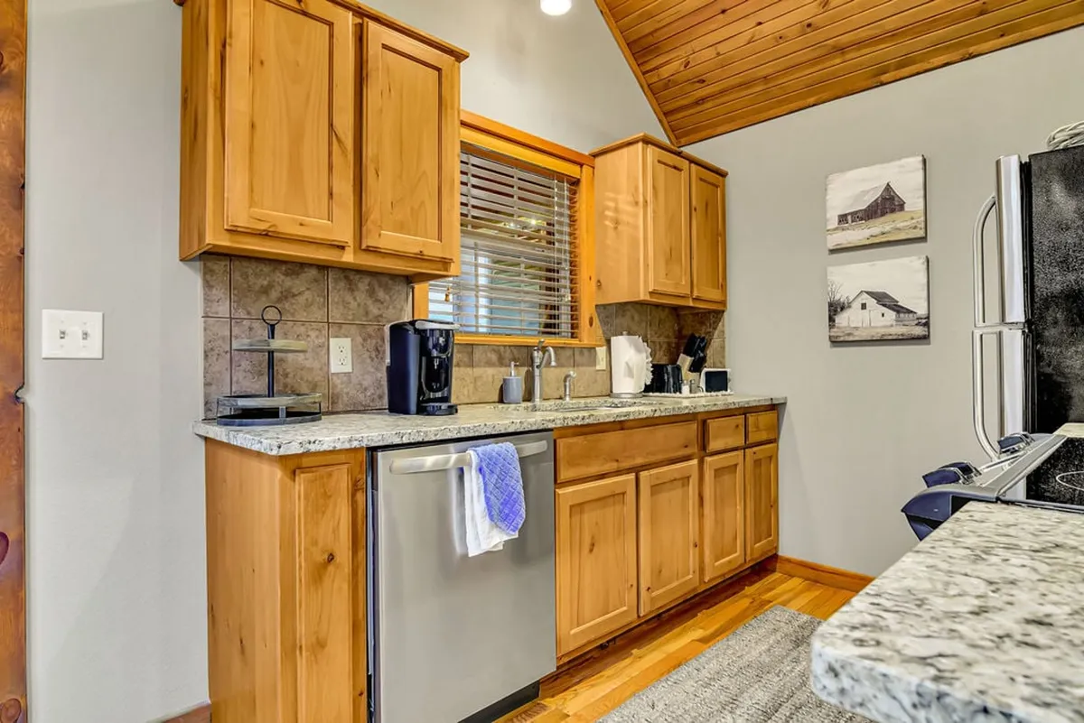 A cozy kitchen interior is depicted with a granite countertop, wooden cabinets, a coffee maker (potentially a Keurig), and a sink. The space is styled for both cooking and socializing, with daylight streaming in to enhance the welcoming mood.