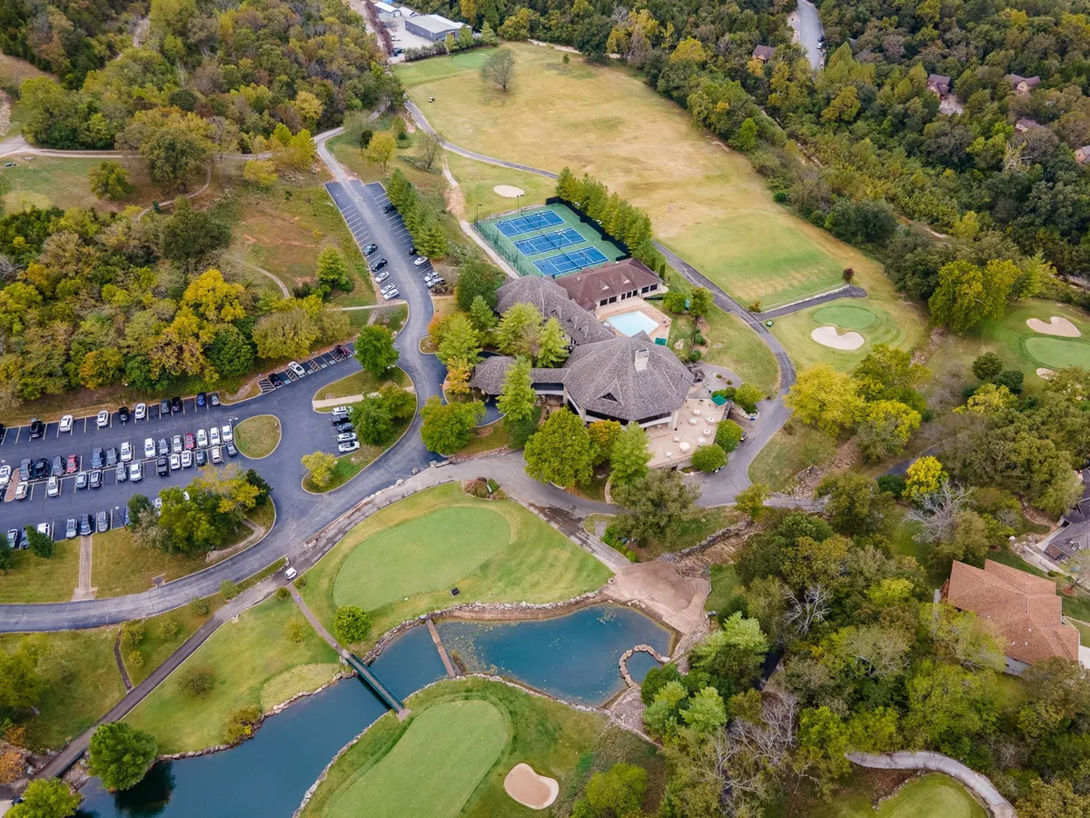 This exterior image captures a sweeping view of the Ledgestone Golf Course, with tennis courts, the clubhouse, a swimming pool, and water features all visible. The lush green landscaping and open sky emphasize the resort amenities and recreational options available within the gated community.