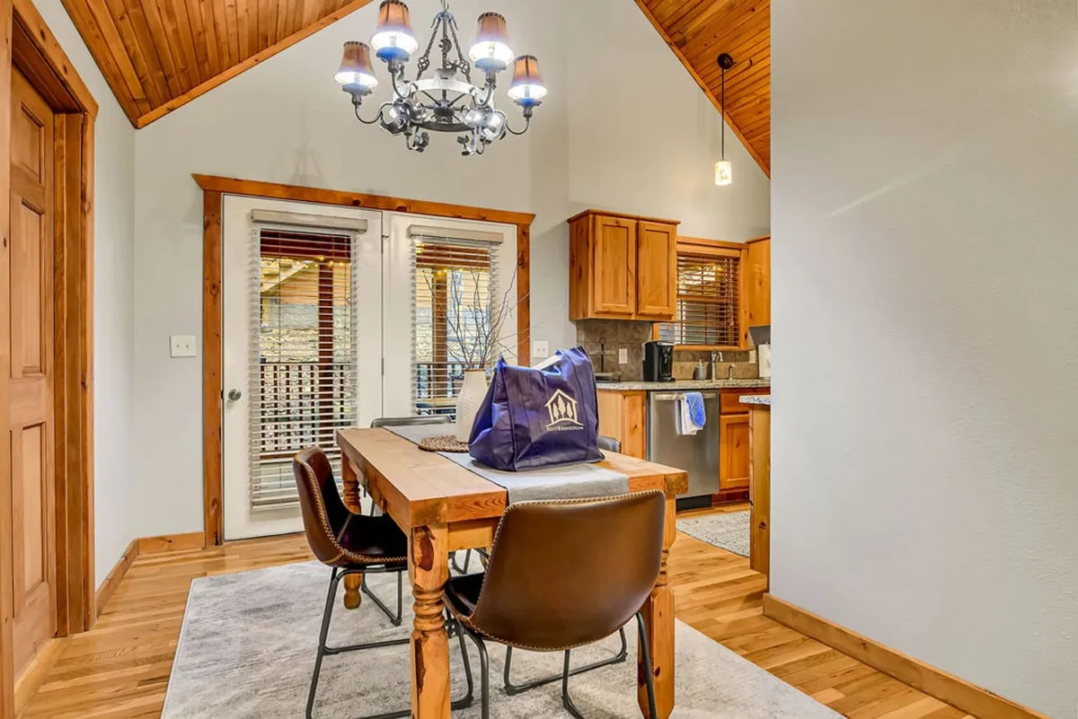 A warmly lit interior highlighting a large wooden dining table under a glowing chandelier, with the kitchen area visible in the background. The inviting setting evokes family dinners, celebrations, and memorable conversations around the table.