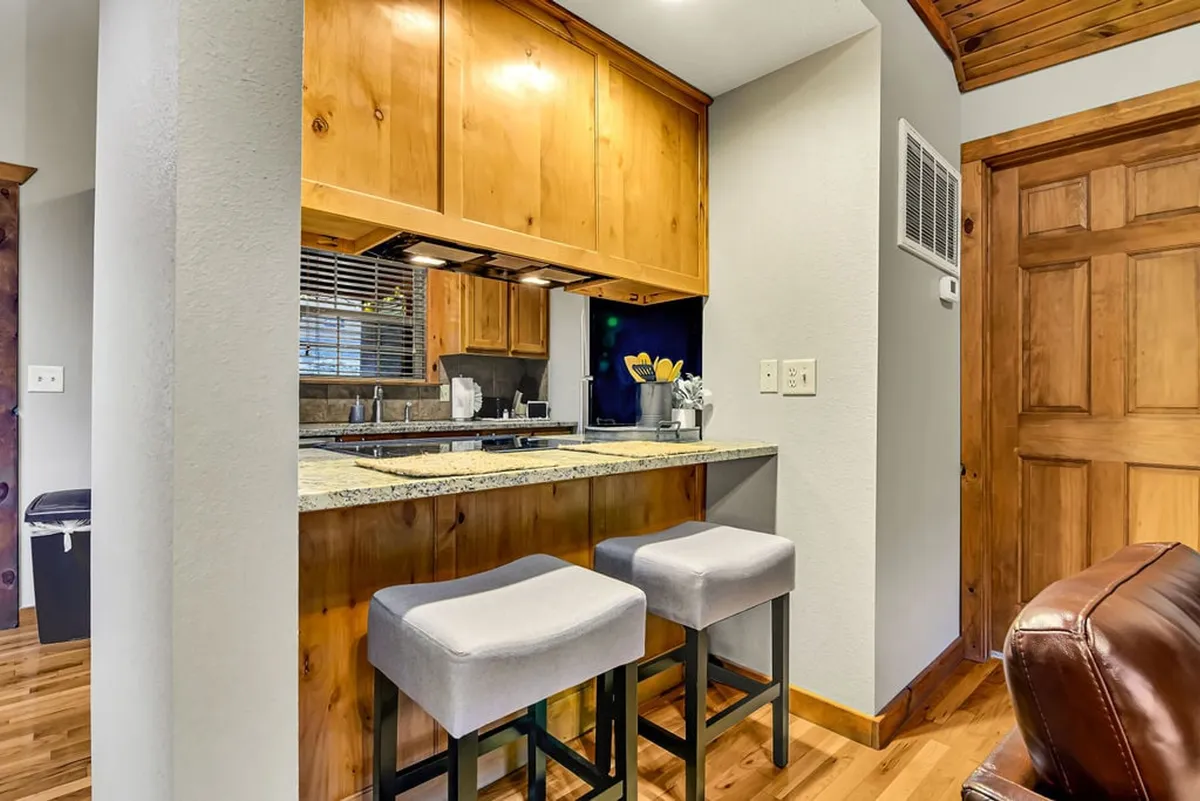 This image features a kitchen with a granite countertop, warm wood cabinetry, and stools at the breakfast bar. Kitchen tools are organized in a holder on the countertop, and the space is well-lit with natural light, creating a cozy and inviting atmosphere for cooking or gathering.