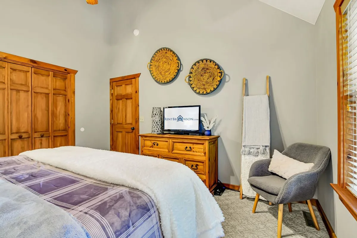 A comfortable bedroom with a bed dressed in soft bedding and decorative pillows, a wooden dresser, TV, and subtle wall art. The space exudes a restful ambiance ideal for unwinding in private.