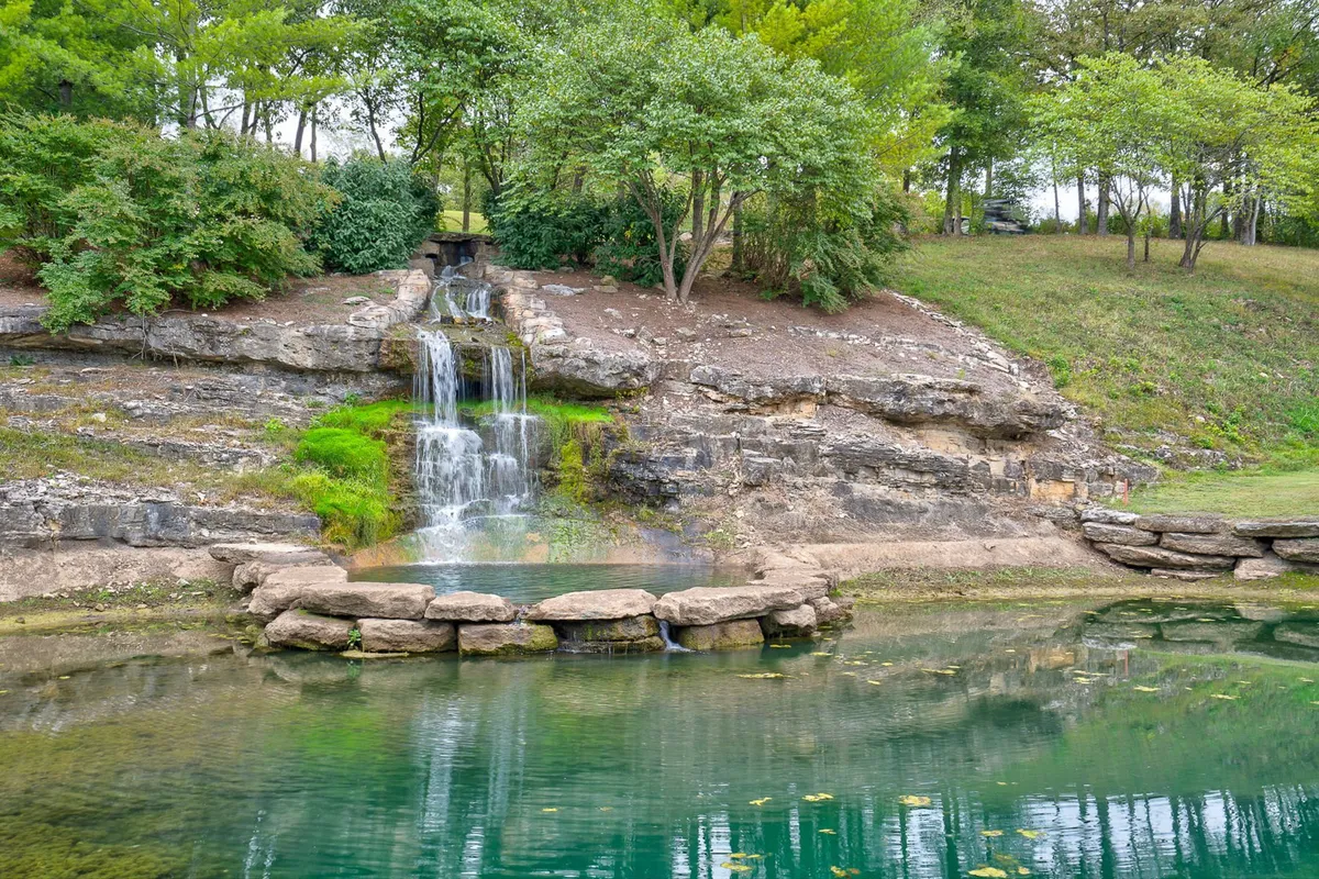 A peaceful pond bordered by rock formations, trees, and a gentle waterfall. The water reflects the surrounding natural beauty, creating a serene environment ideal for fishing, relaxing, or enjoying a quiet walk.