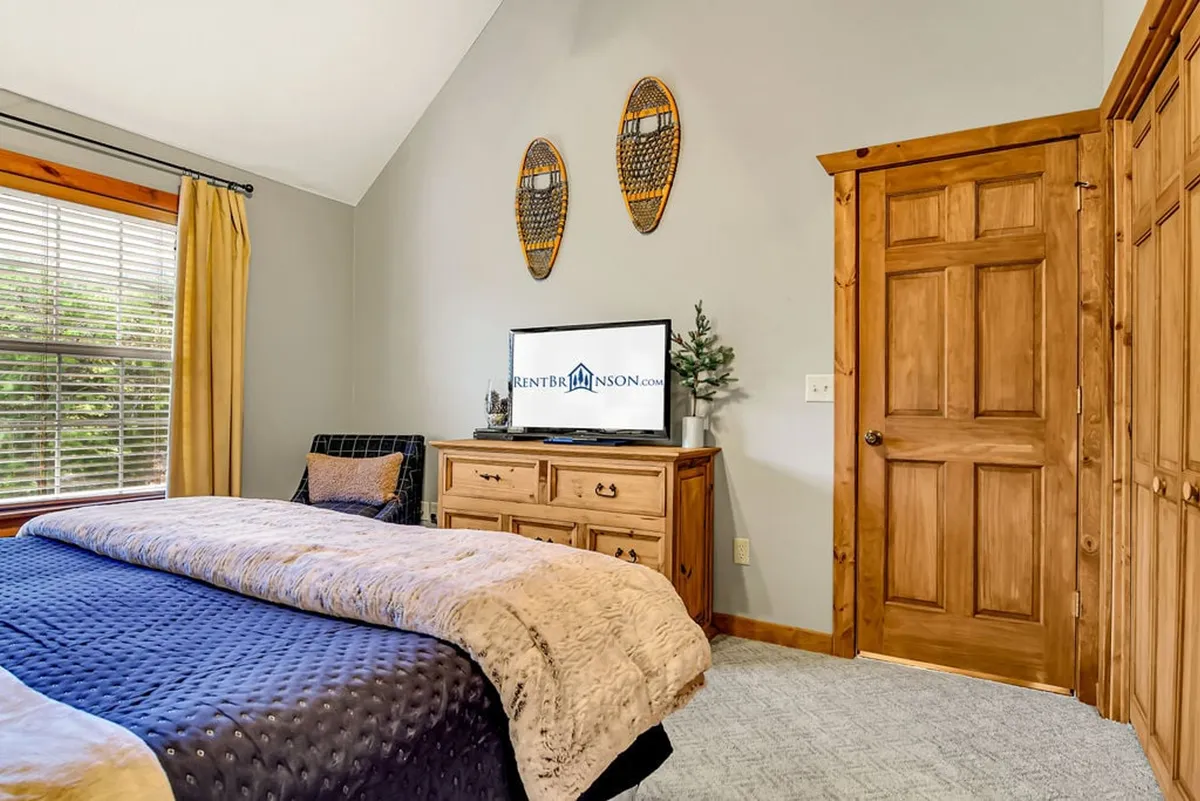 A warmly lit bedroom featuring a plush king bed topped with a soft blanket, a wooden dresser, and a wall-mounted TV. Decorative snowshoes add character, and natural light filters in through a window, enhancing the cozy, restful atmosphere.