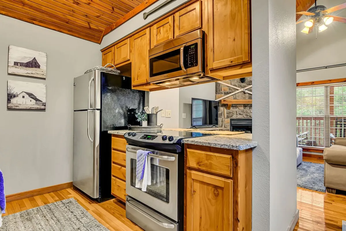 This image presents a kitchen area featuring modern appliances, wooden cabinets, a stone fireplace, and a window letting in natural light. The setting is ideal for cooking, entertaining, and relaxing while staying connected to adjacent living spaces.