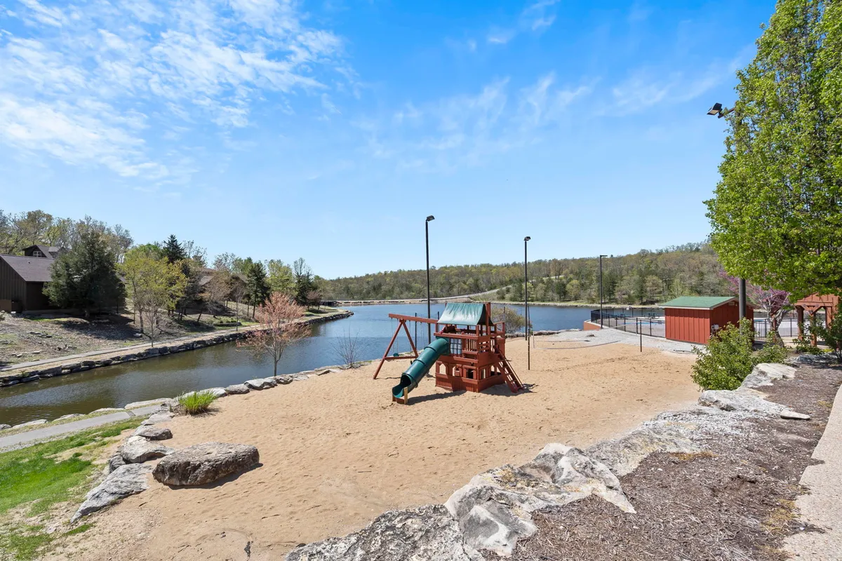 A joyful exterior scene showcasing vibrant playground equipment set in a sandy area along a picturesque waterfront, surrounded by lush green trees and rock formations. Children are actively playing while families gather nearby, capturing the essence of outdoor fun and family bonding.