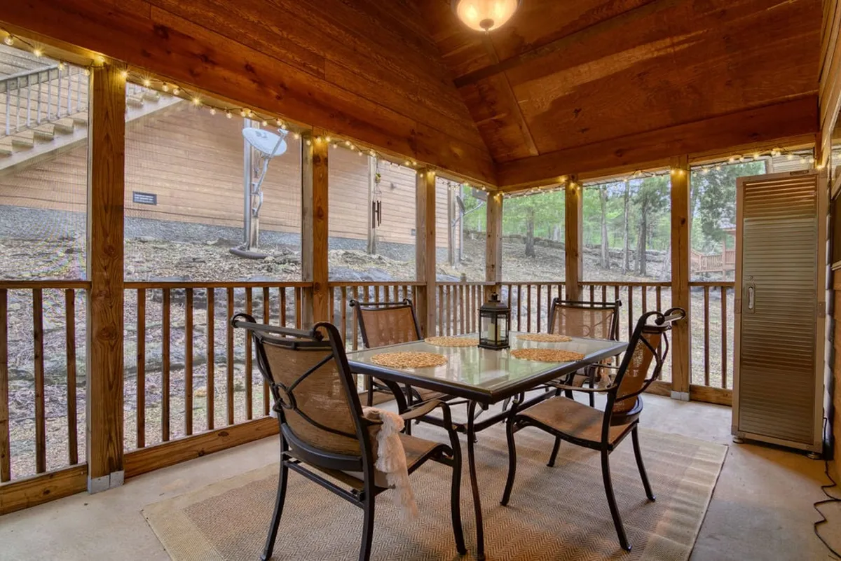 This image captures a cozy screened-in porch featuring a dining table, hanging light fixture, and views through screened walls to the outdoors. The communal table and soft lighting evoke a sense of relaxed gatherings and meals enjoyed in the fresh air, with nature visible just beyond the screens.